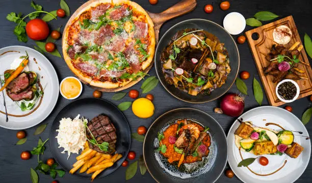 Assorted gourmet meals on a dark surface; includes arugula-topped pizza, steak with fries, beef medallions, pasta in sauce, tofu skewers, and seasoned meat on a board, surrounded by fresh herbs and tomatoes.