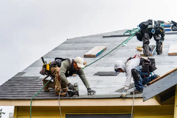 Roof construction workers in safety gear installing shingles on a house